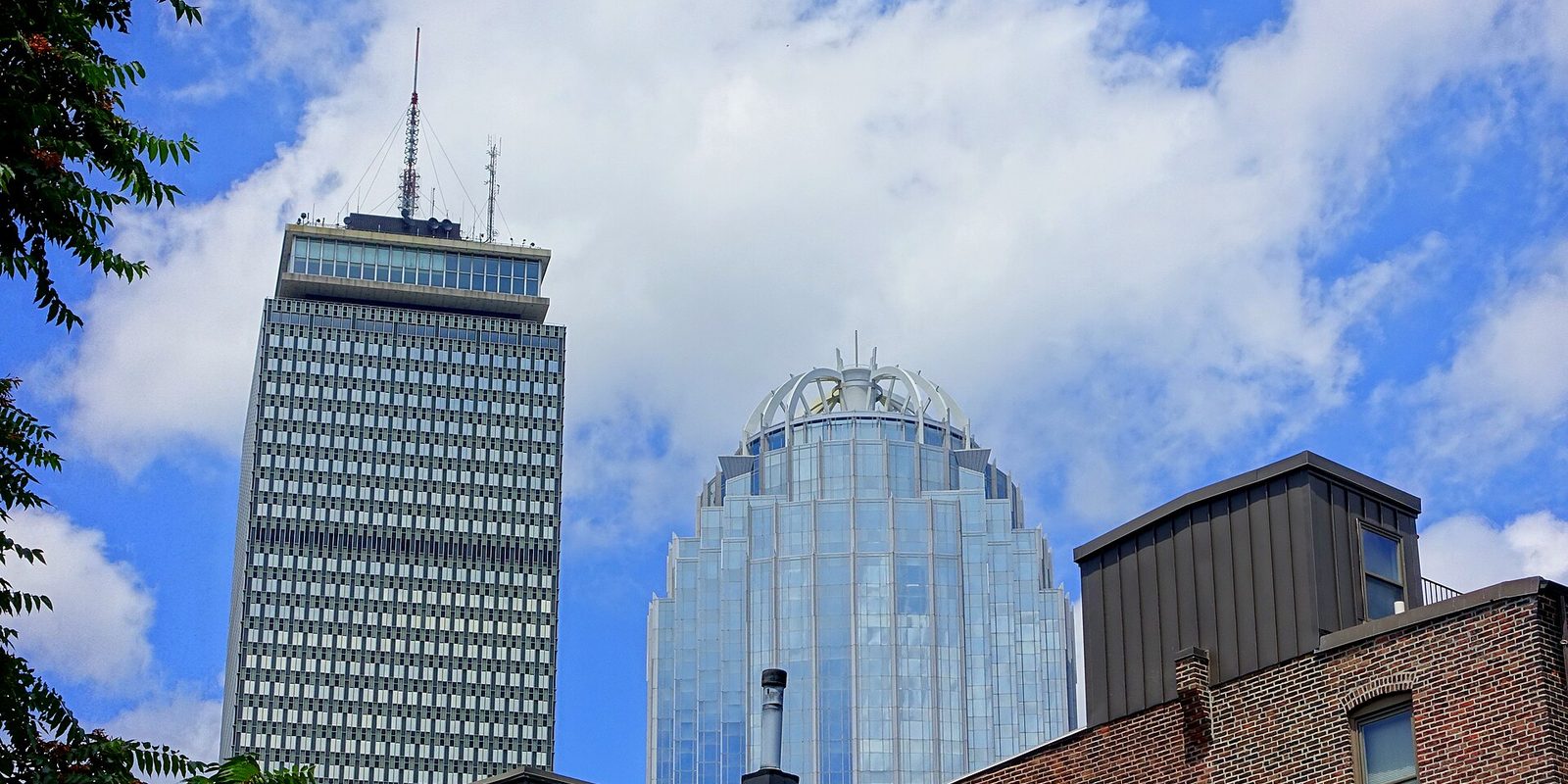 Boston towers seen from street level, a tall antenna-topped skyscraper and a glass tower with a crowned dome rising past a brick rooftop into a bright sky