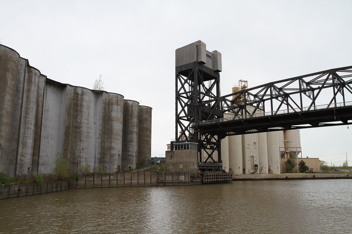 Concrete grain elevators line the Buffalo River beside a black steel vertical-lift railroad bridge under a flat gray overcast sky.