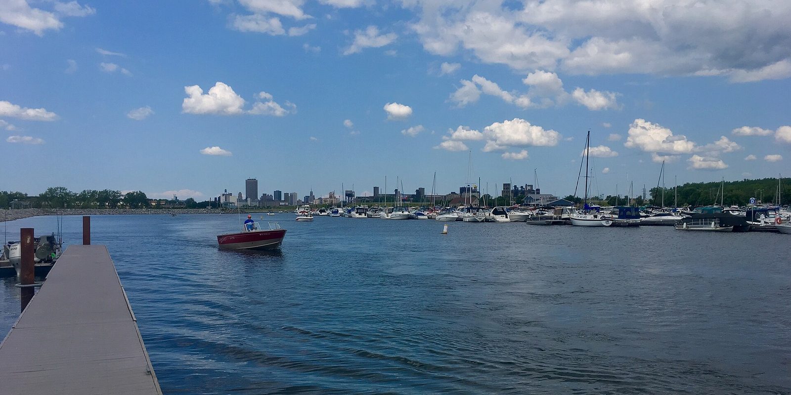 Buffalo's waterfront from a marina dock, a small red motorboat passing moored sailboats with the low downtown skyline across the water