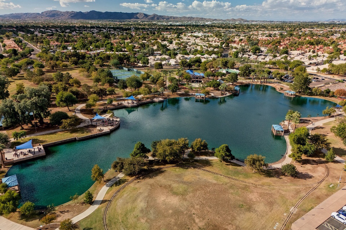 A Chandler park from above, a bright blue lake with wooded islands set in mown grass and paths, houses and mature trees around it and desert mountains on the far horizon.