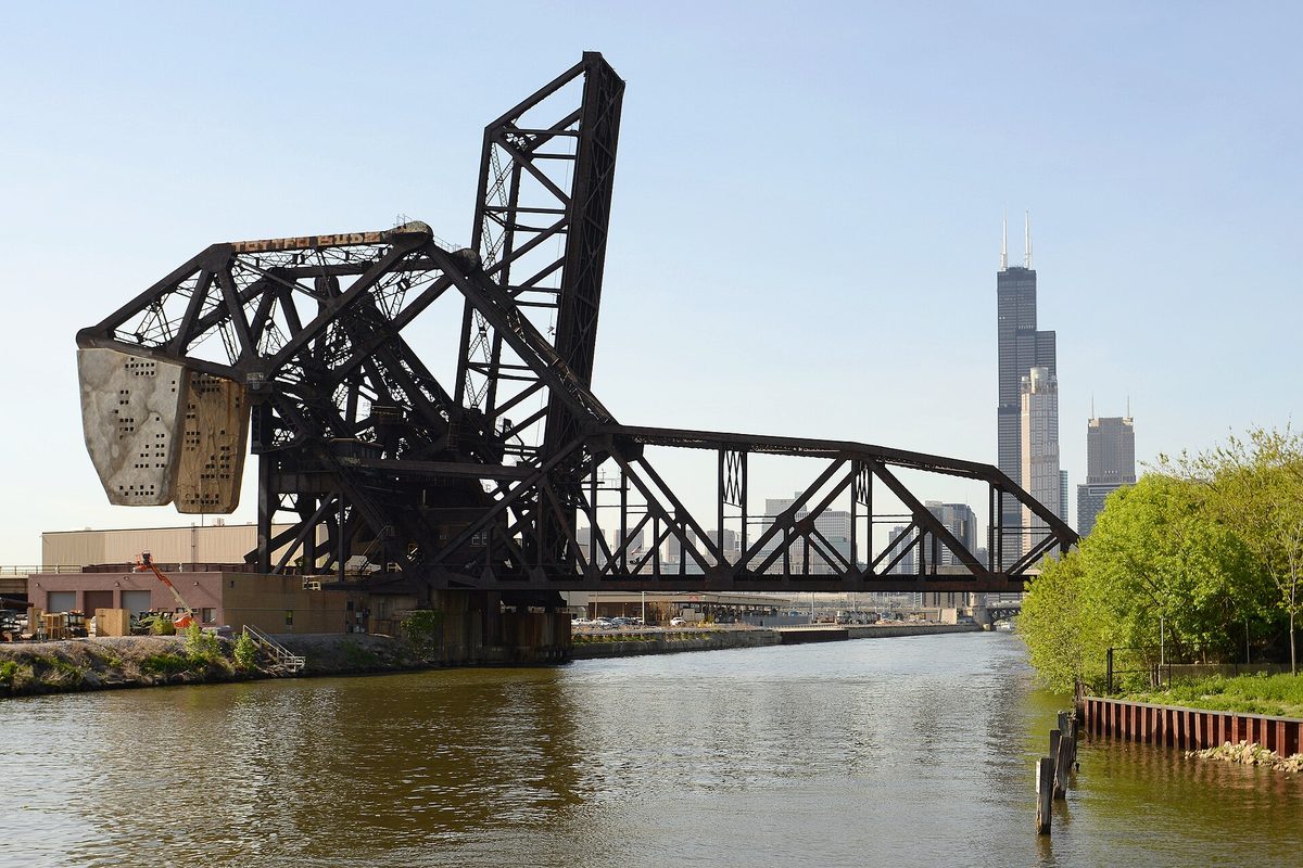 A rusted railroad bascule bridge stands raised over the Chicago River, with the Willis Tower and the downtown skyline behind it.