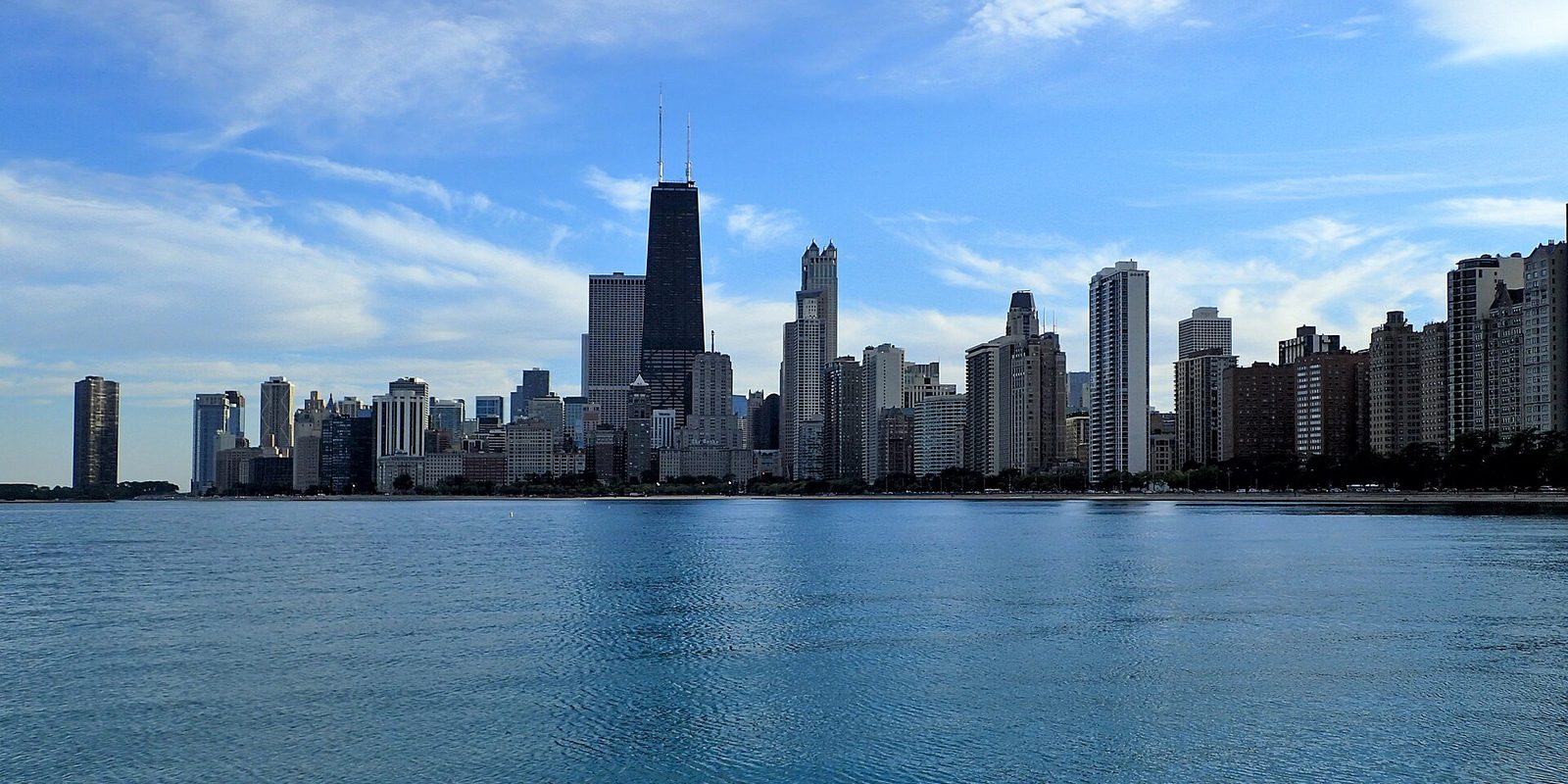 Chicago's lakefront skyline across calm blue water, a dark tapered tower with twin antennas standing above the surrounding high-rises