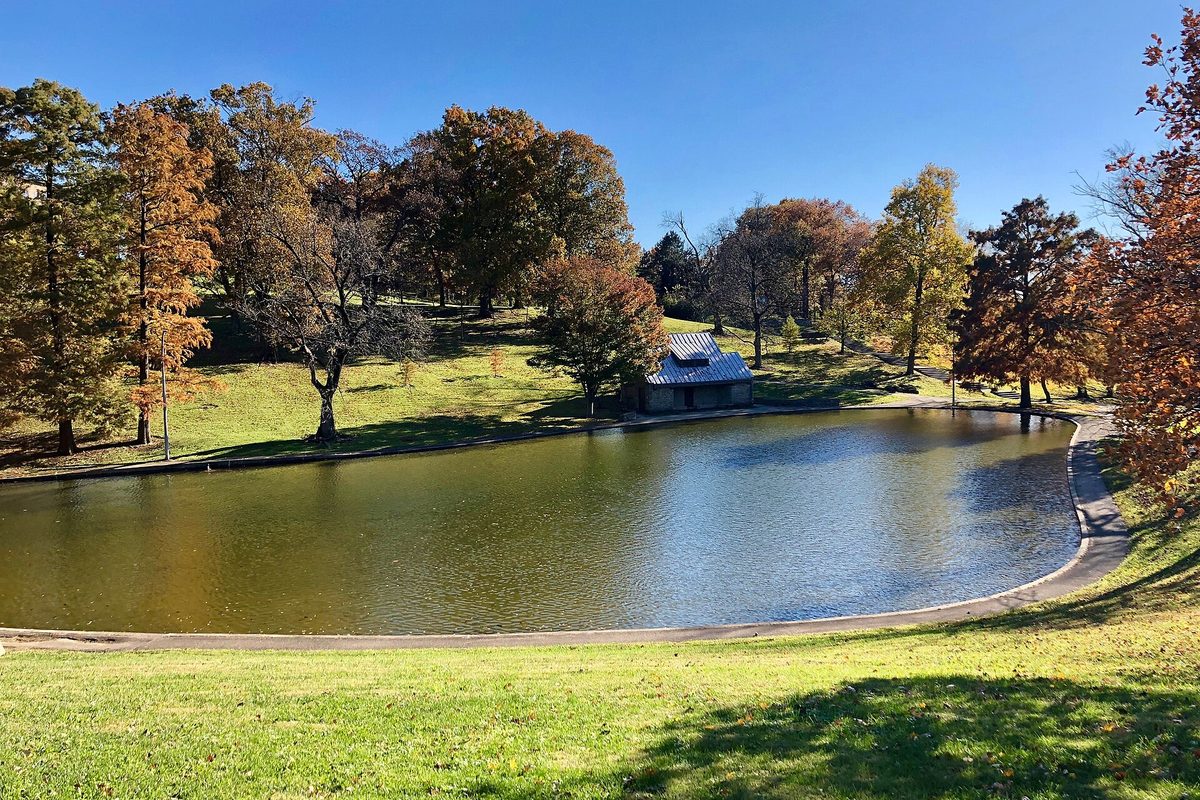 Cincinnati park pond ringed by a paved walking path, with a small stone shelter and orange autumn trees under a clear blue sky.