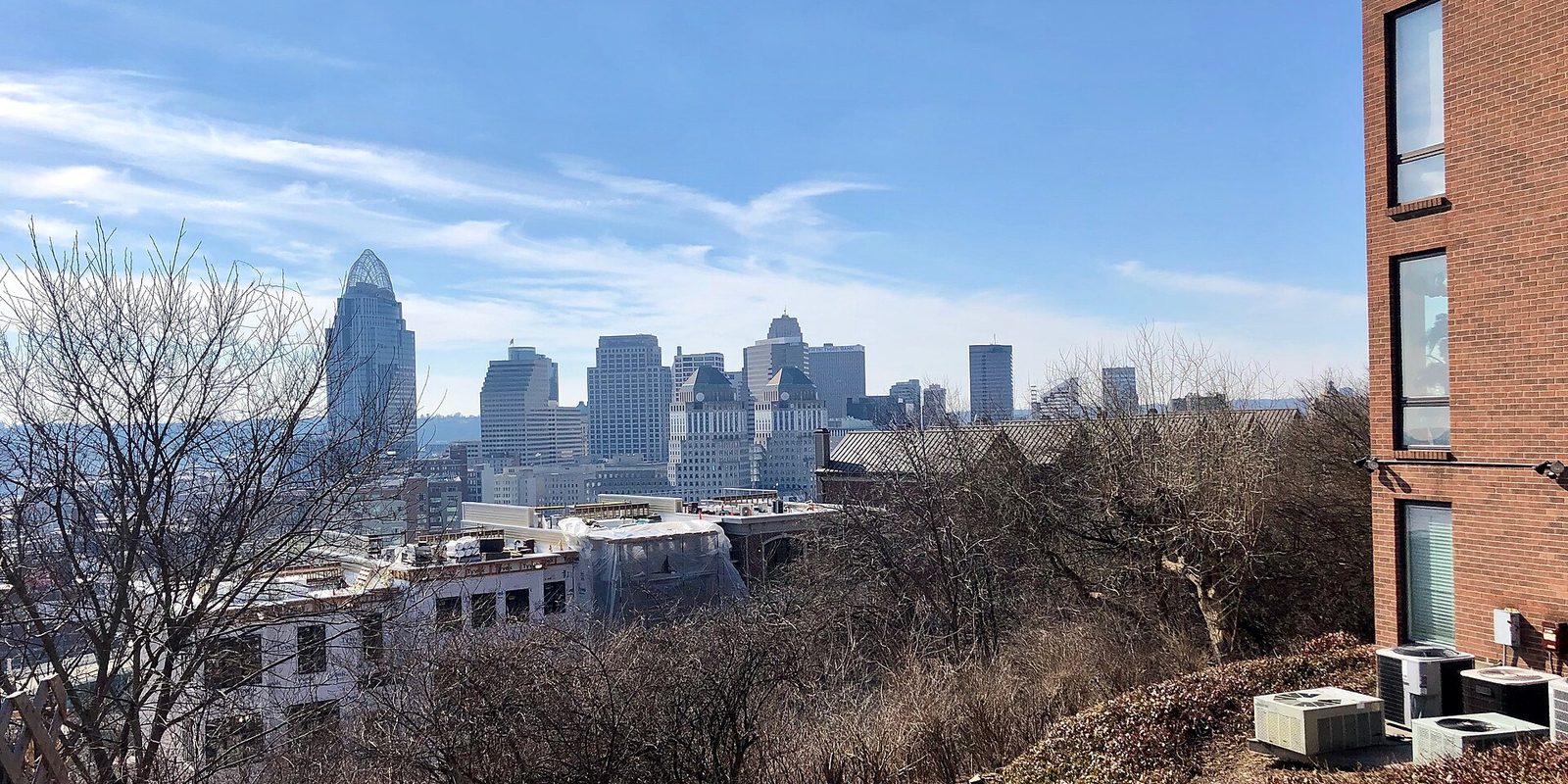 Cincinnati's downtown towers seen from a wooded hillside over bare grey trees and rooftops, a brick apartment block at the right, under a clear blue sky