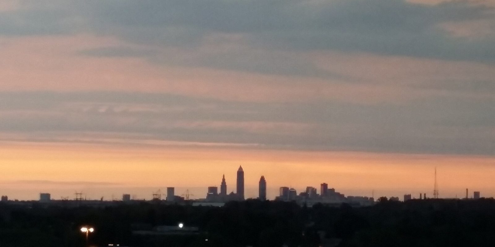 Cleveland's downtown towers standing as a distant silhouette above a dark treeline, under a wide pink and orange sunset sky