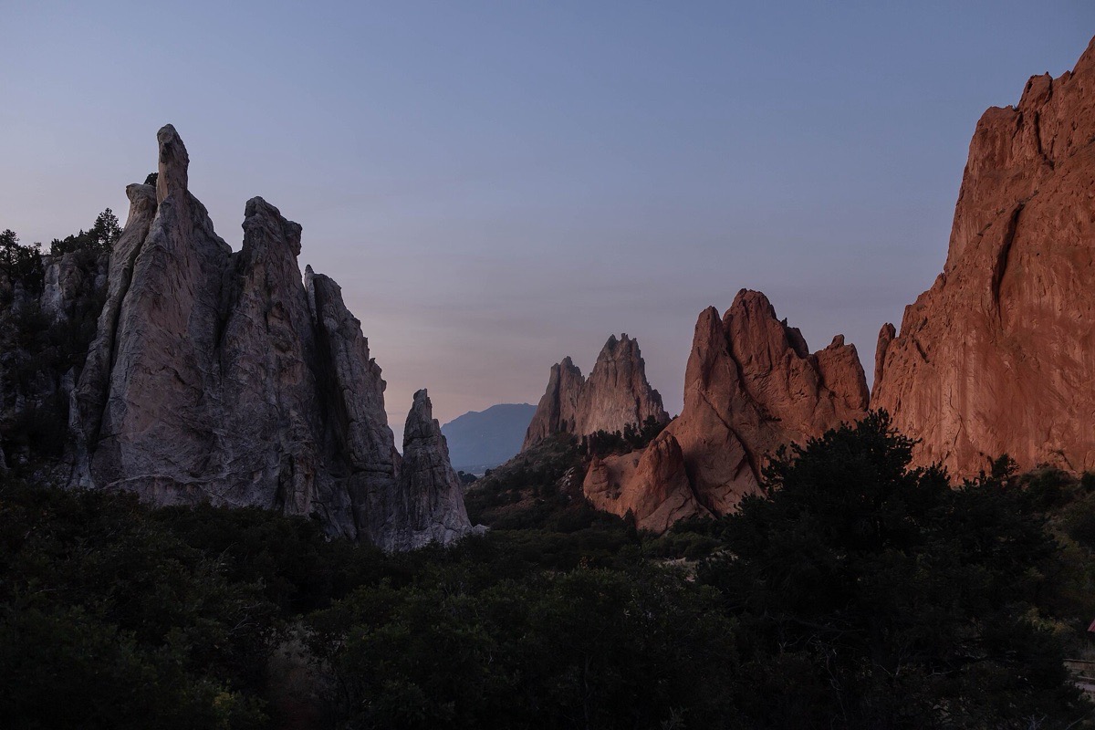 Tall red and grey sandstone fins standing above dark conifers at sunrise in Colorado Springs, the nearest one pale against a soft violet sky with a mountain visible in the gap beyond.