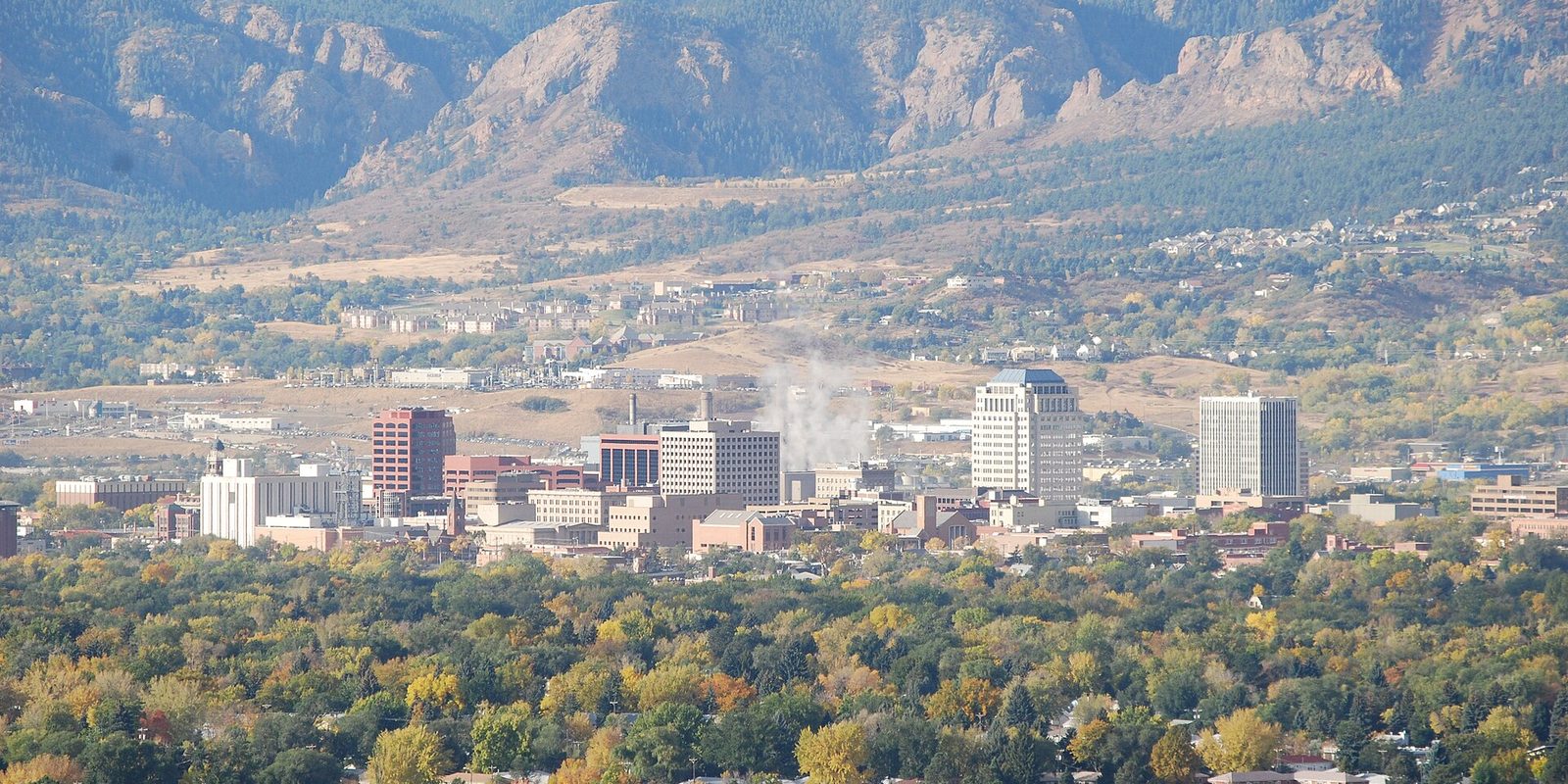 Colorado Springs downtown viewed from above across a canopy of green and yellow autumn trees, with rocky foothills rising behind the city