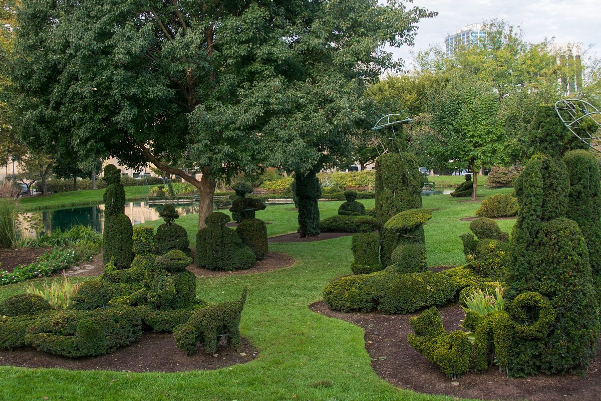 Columbus topiary garden where clipped shrubs form human figures and a dog on the lawn beside a pond, a tower rising past the trees.