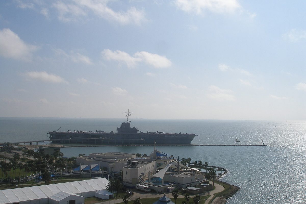 A retired aircraft carrier moored in Corpus Christi Bay with planes on its flight deck, waterfront buildings and palms below