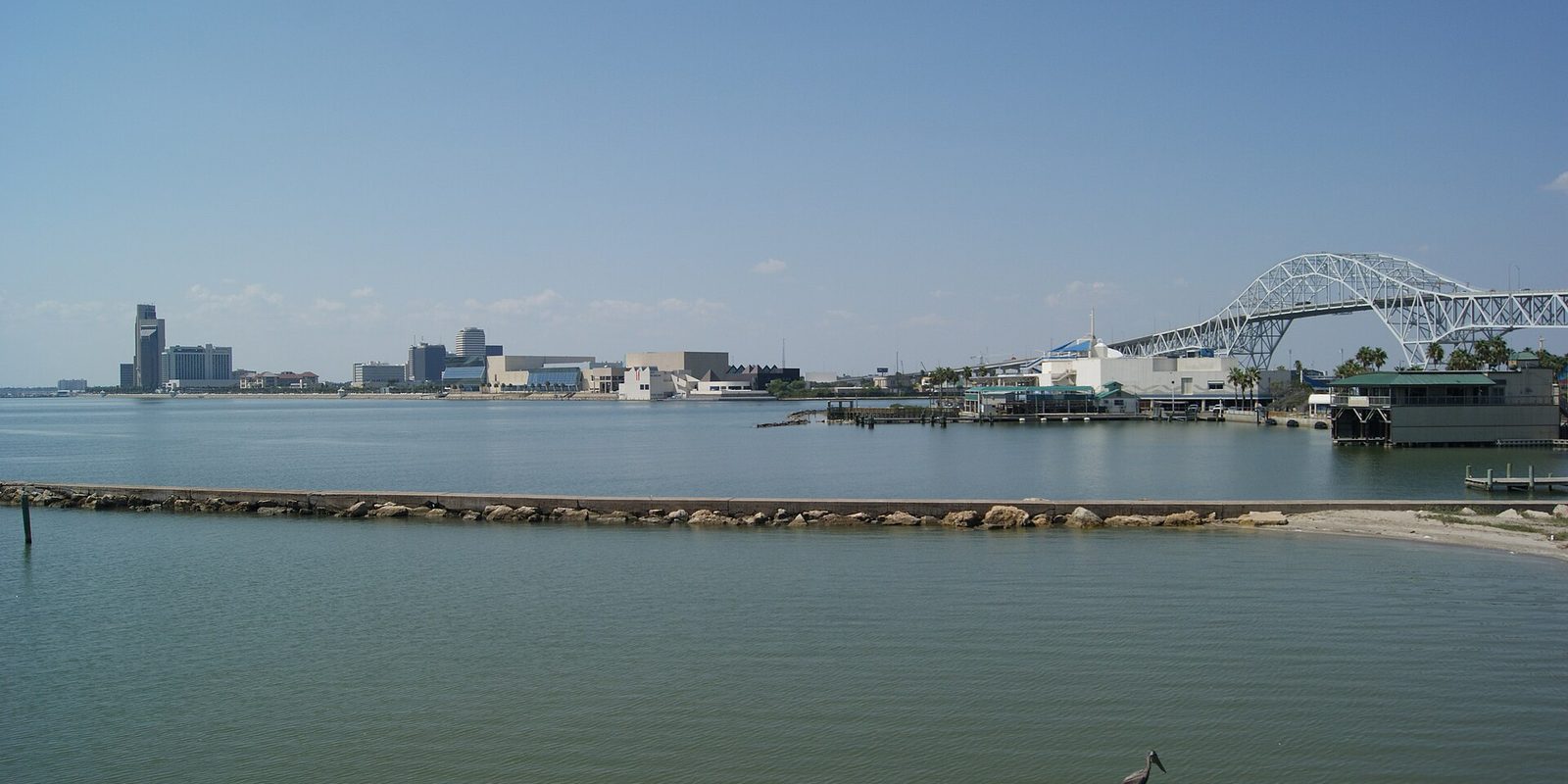 Corpus Christi seen across calm bay water, with low downtown buildings and waterfront halls on the left and a tall steel arch bridge on the right