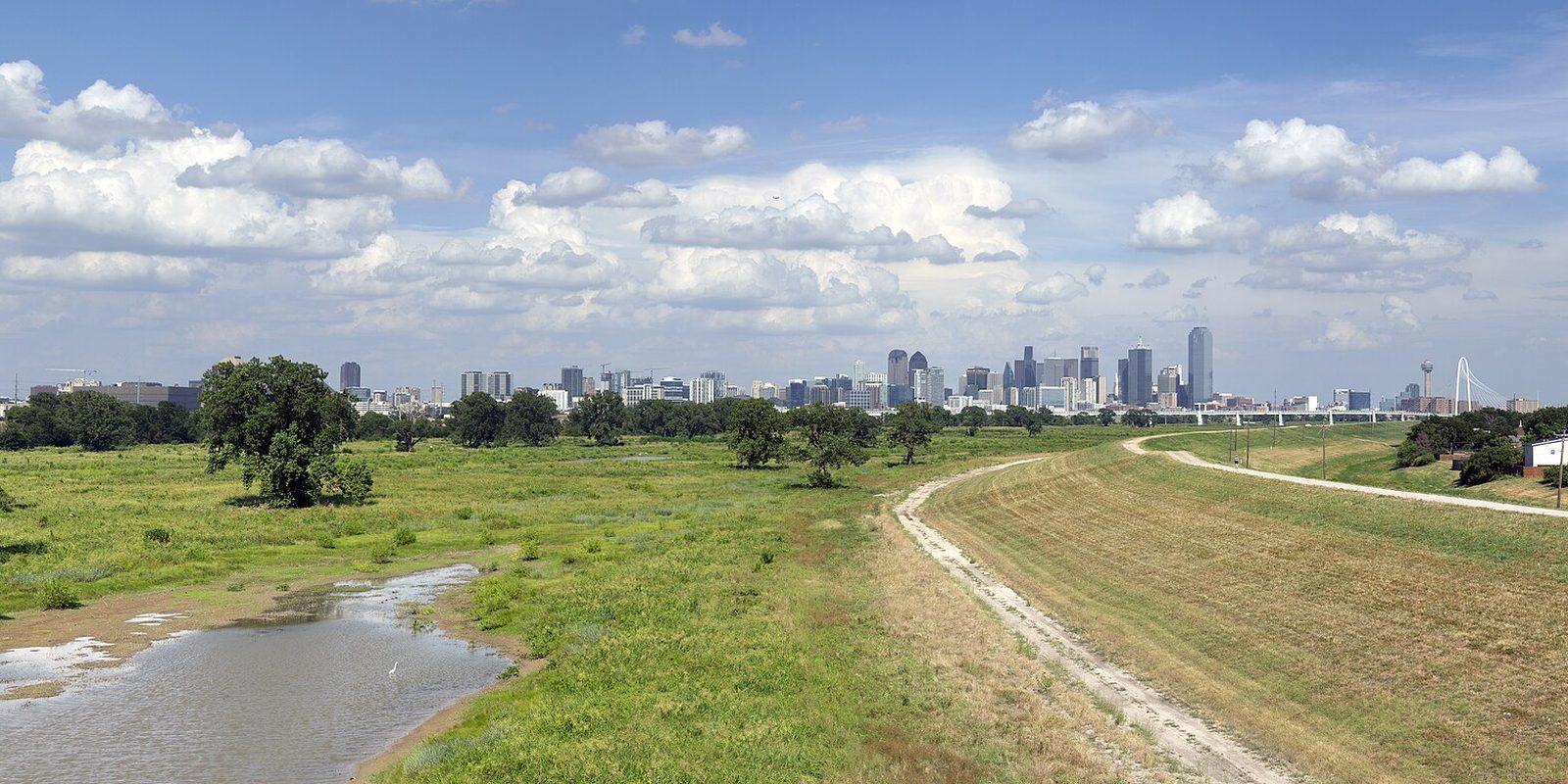 Dallas skyline low on the horizon beyond a wide green floodplain of grass, trees and shallow standing water, with a dirt levee track and tall cumulus clouds