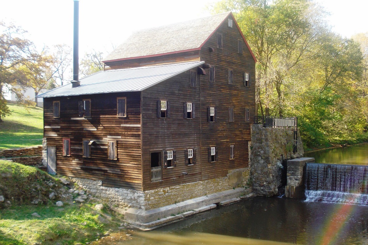 A tall timber grist mill standing on a stone foundation beside a low dam in Iowa, water spilling over the sill and autumn trees crowding the bank behind.