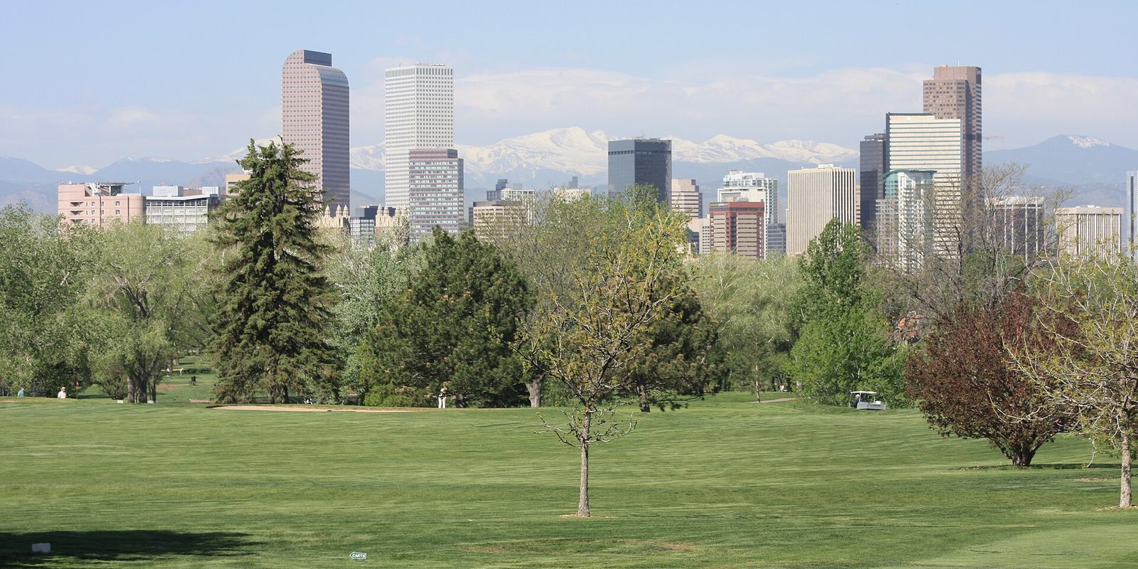 Denver skyline rising behind a broad mown green and leafy trees, with the snow-capped Rocky Mountains stretched across the hazy horizon