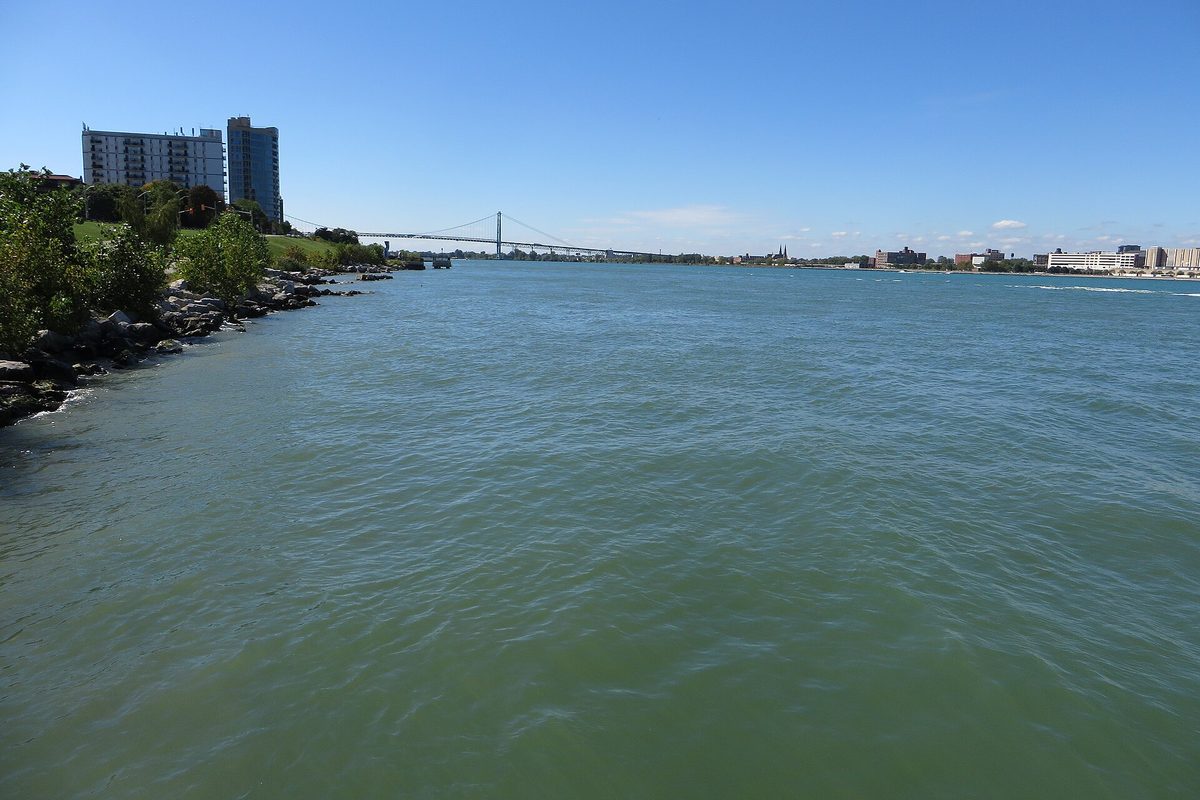 The Detroit River runs green-blue past a rocky shoreline toward a suspension bridge and the distant downtown skyline under a clear sky.