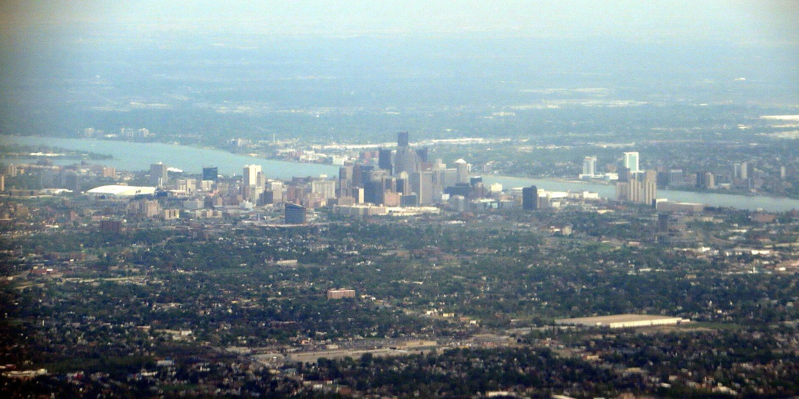 Hazy aerial view over Detroit, downtown towers clustered along the river with the far bank beyond and mile after mile of tree-filled neighborhoods in front
