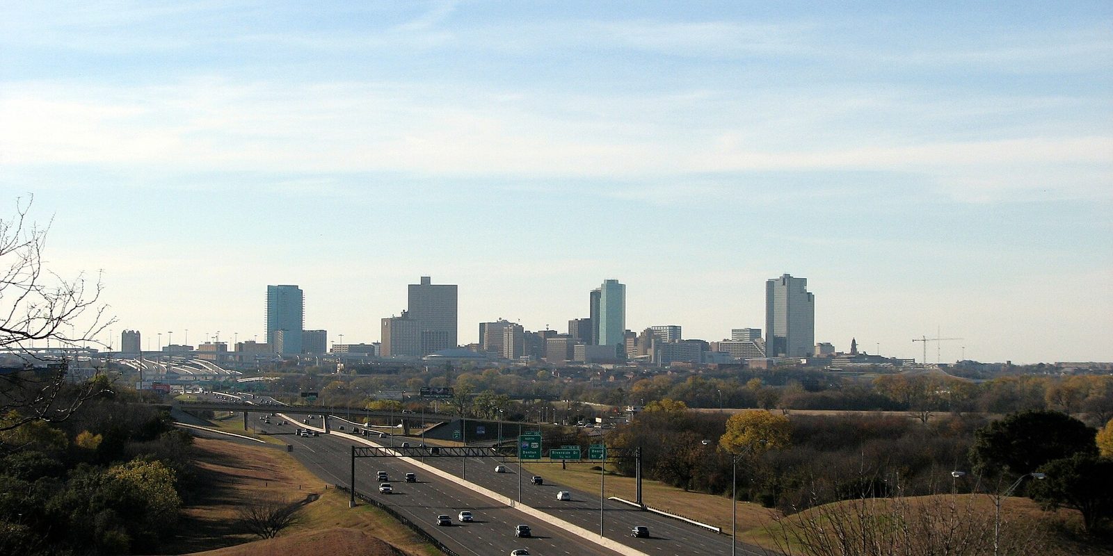 Fort Worth skyline seen from a rise across a wide multi-lane freeway, with bare branches and dry tan grass in the foreground under a pale sky