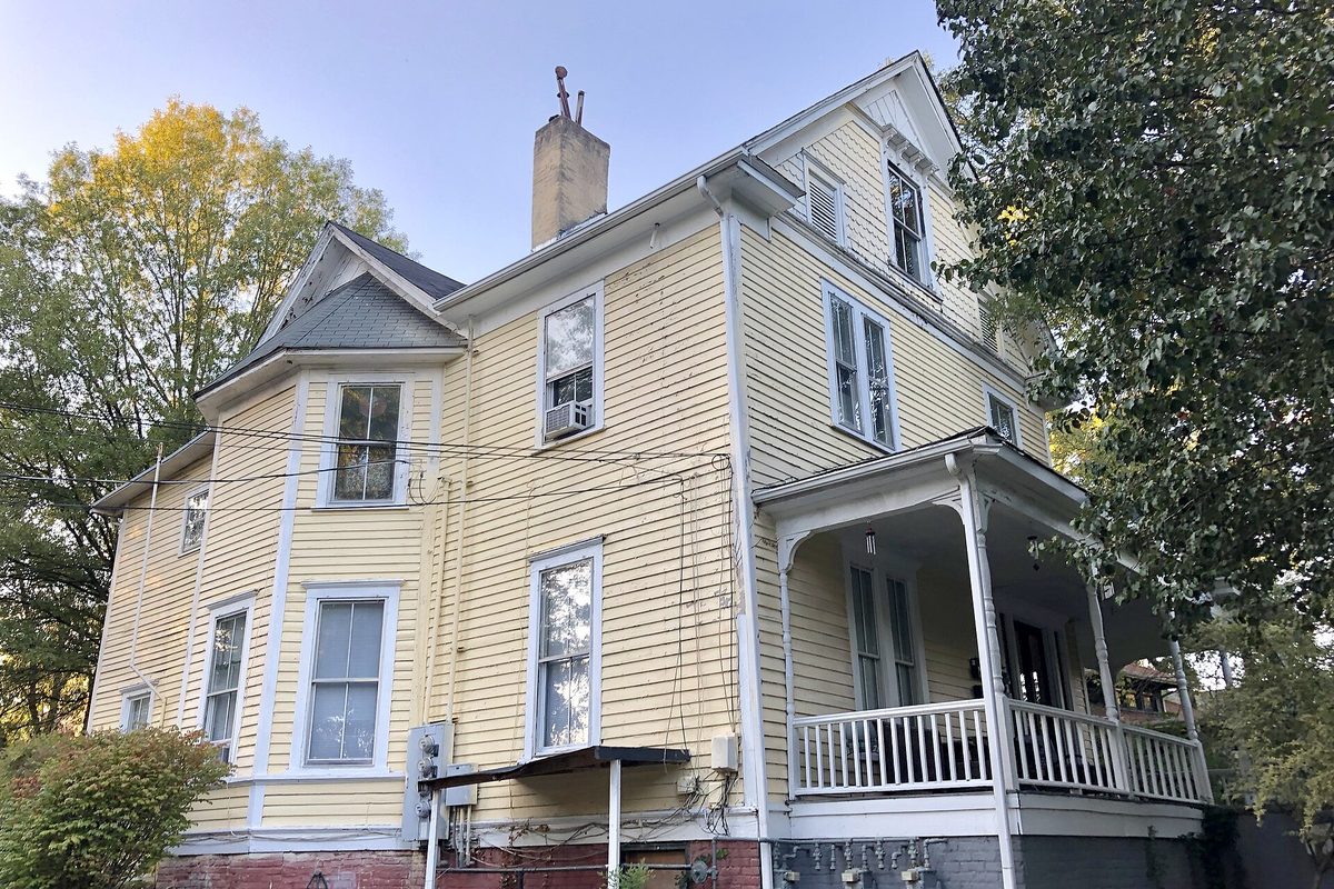 A two-story yellow clapboard Greensboro house with white trim, a columned front porch and tall brick chimney, lit by low golden light.