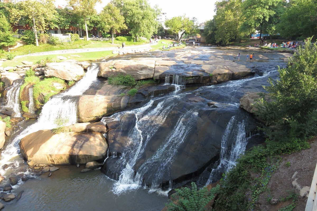 A South Carolina river dropping in white sheets over broad tilted rock shelves in the middle of a city park, green lawn and mature trees on the banks above.