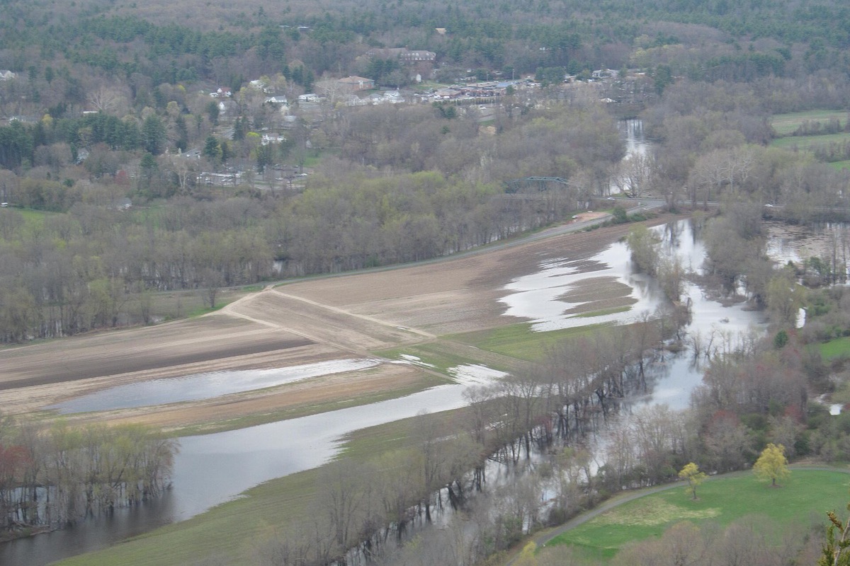The Connecticut valley seen from a ridge, the river spread out in flood across pale sandbars and green fields with a wooded village on the far side.