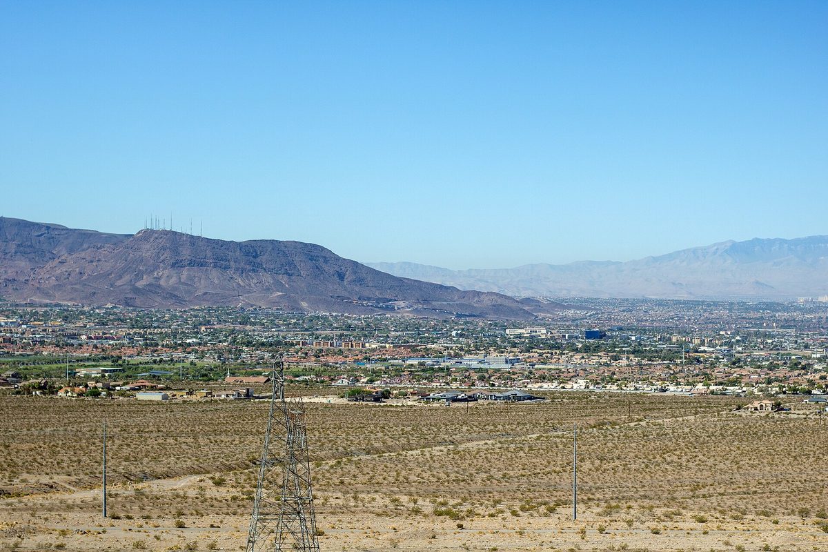 Henderson's tile-roofed subdivisions spread below an antenna-topped desert ridge, with a steel transmission tower rising from scrub in the foreground.