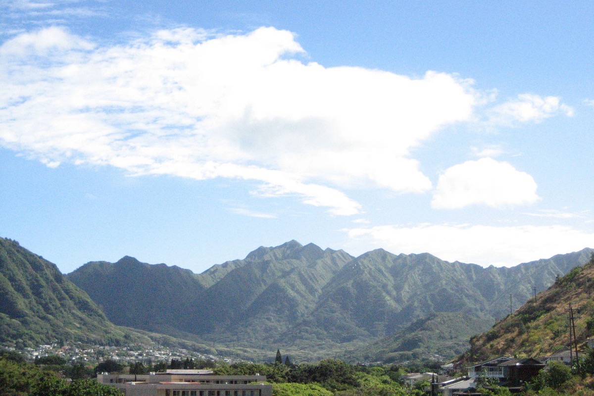 The deeply folded green ridges of a valley behind Honolulu rising into cloud, houses and low buildings scattered across the flat valley floor beneath them.