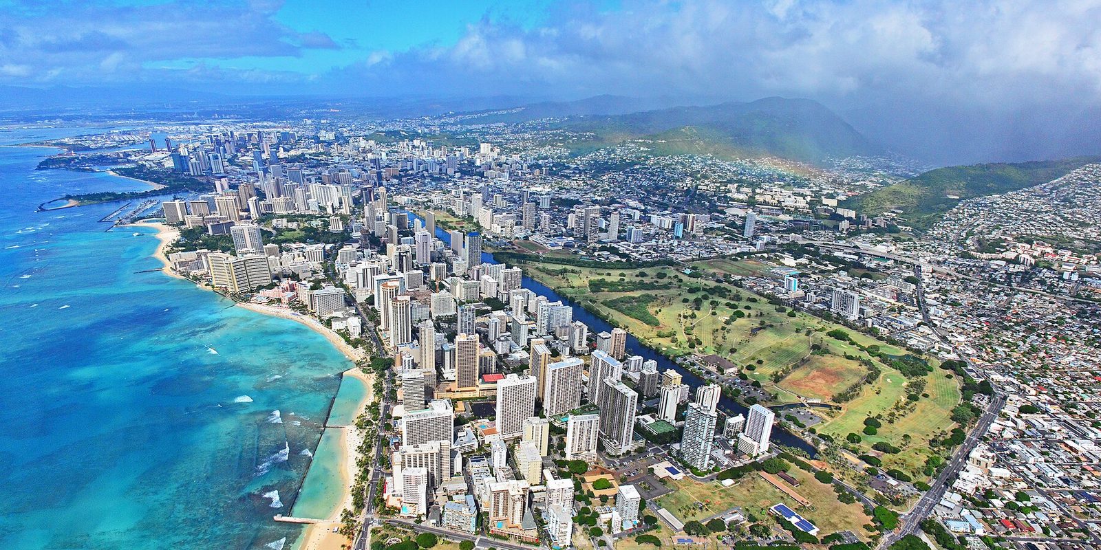Aerial view of Honolulu with turquoise reef water and beach beside dense Waikiki high-rises, green ridges and parkland inland under scattered clouds