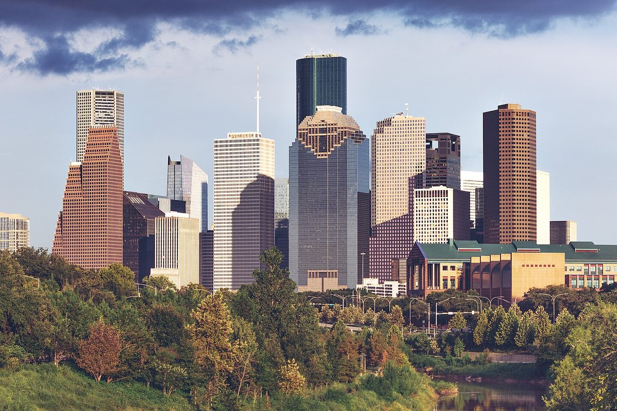 Houston's downtown towers catch warm low sunlight beneath dark clouds, above a wooded bayou bank and a sliver of brown water.