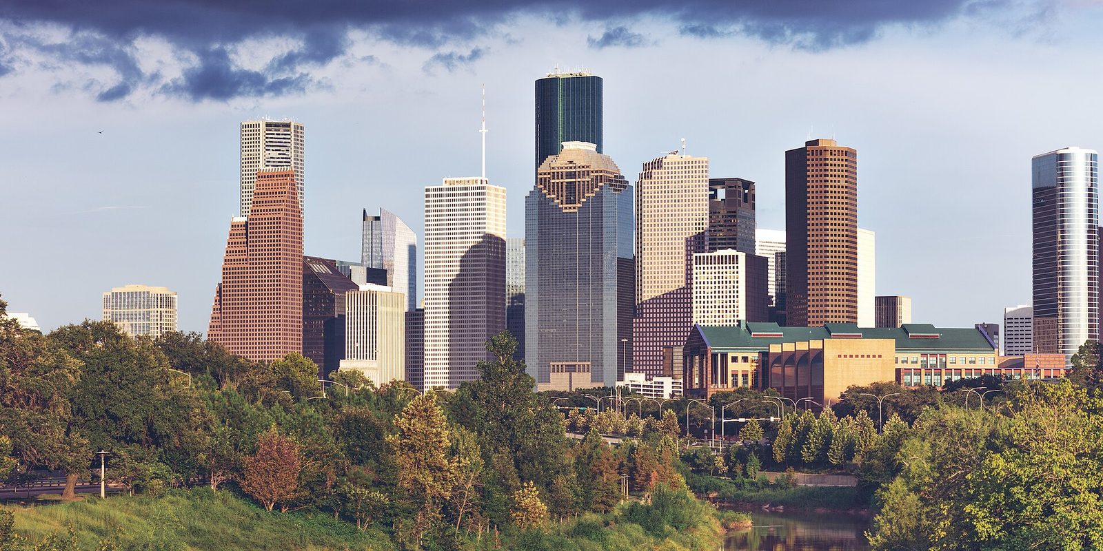 Houston's downtown towers rising behind a thick belt of green trees and a sliver of bayou, lit warm and low with dark clouds over a blue sky