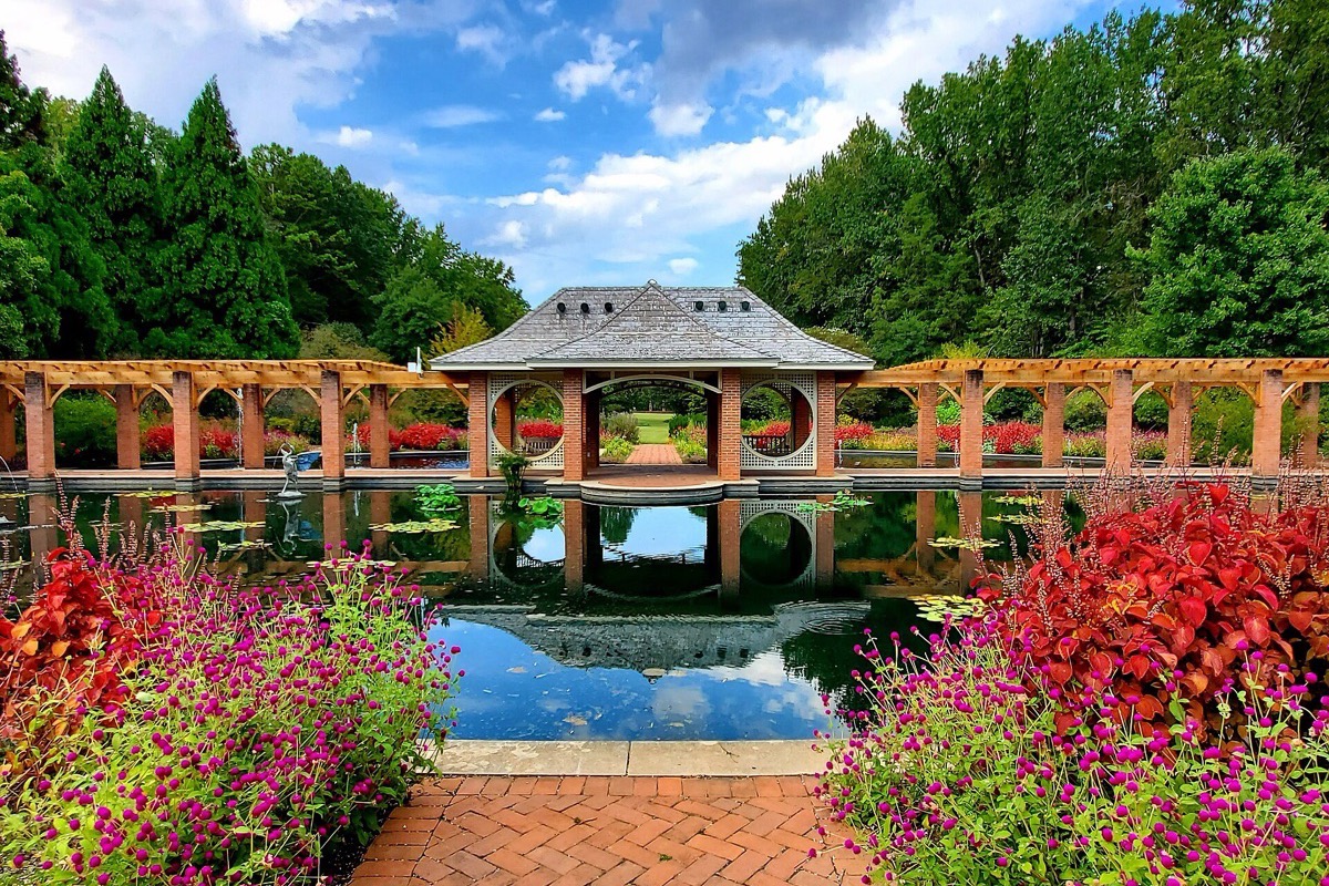 A formal Alabama water garden, a timber pavilion reflected in a still rectangular pool with beds of red and pink flowers along a brick terrace in front.