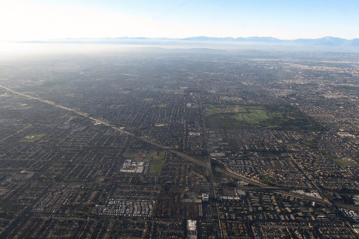 Aerial over Irvine's flat street grid of tract housing and low-rise blocks, a freeway cutting diagonally toward hazy mountains on the horizon.
