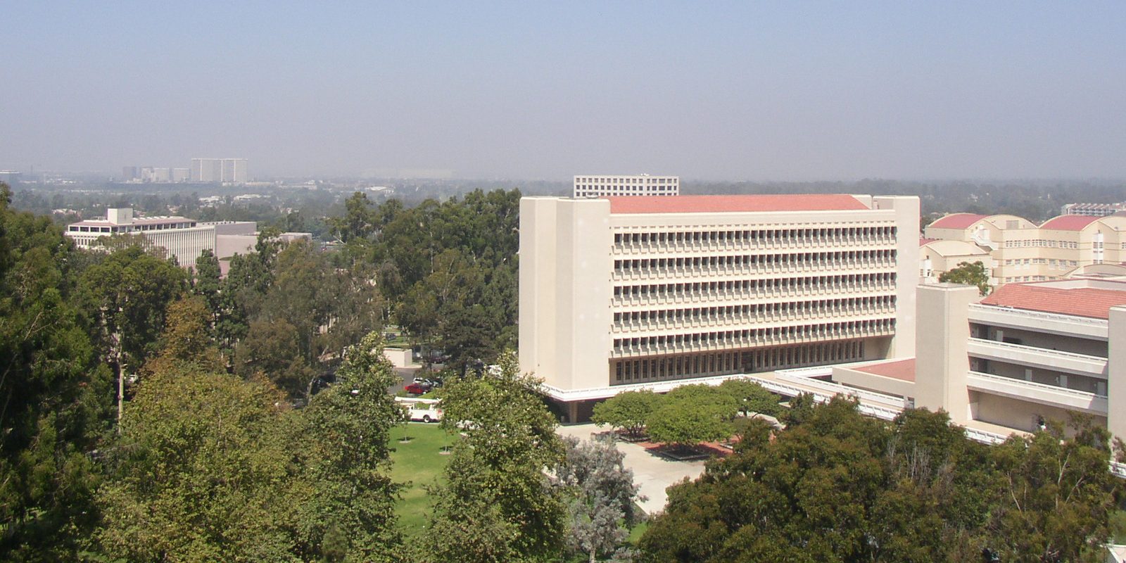 Elevated view over Irvine, California, with a long cream campus building under a red tile roof among mature trees and hazy low towers on the horizon