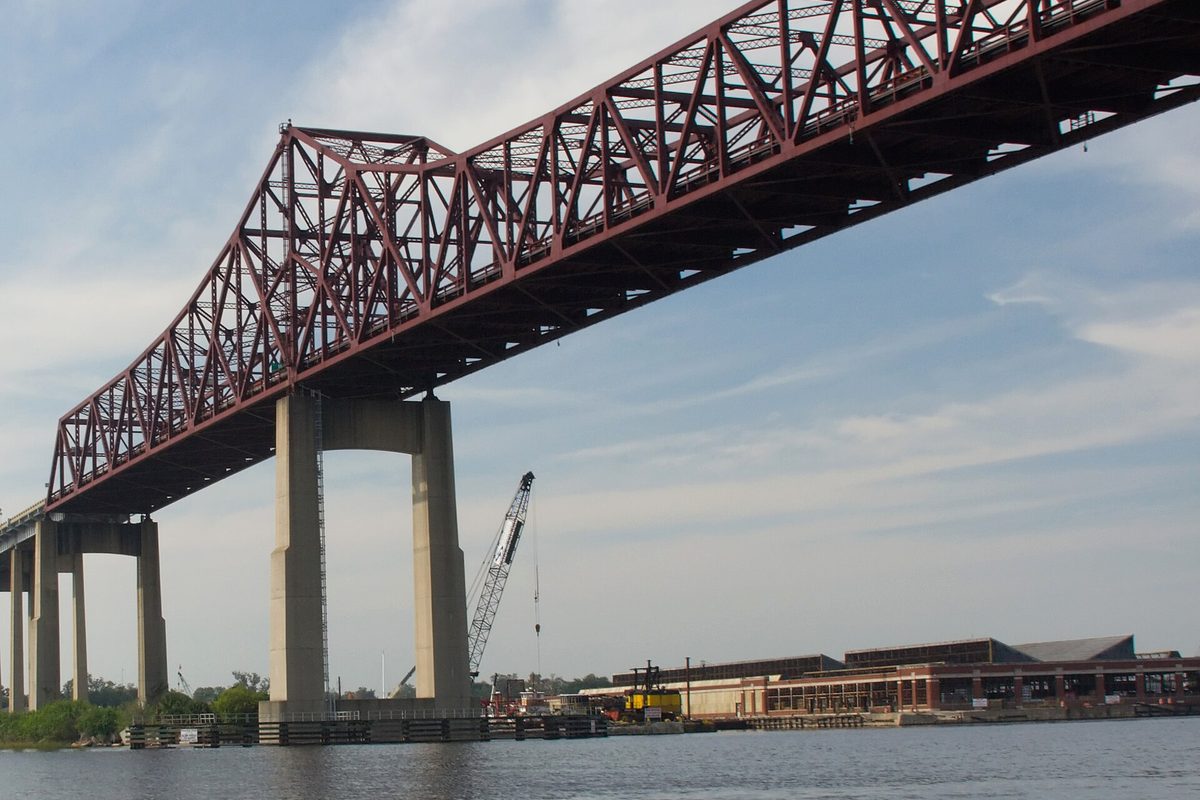 A dark red steel truss bridge on tall concrete piers crosses a wide river in Jacksonville, past a dockside warehouse and a lattice crawler crane.