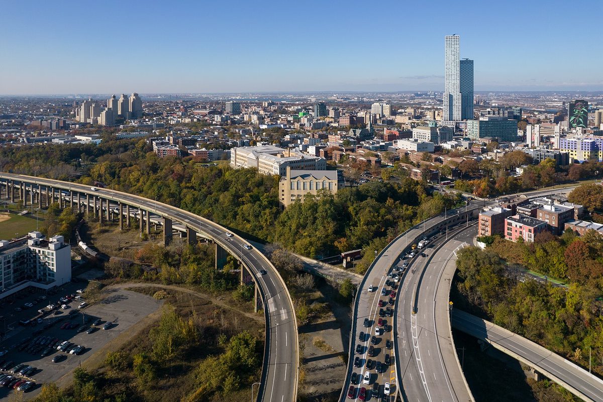 Jersey City, NJ: elevated highway viaducts curve past autumn treetops toward a downtown skyline topped by a tall glass tower under clear blue sky.