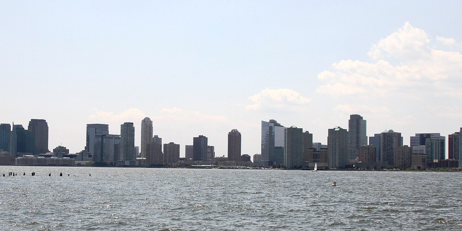 Jersey City's waterfront towers seen across the Hudson River, a lone sailboat near shore and sunlight glinting on the water under a hazy blue sky