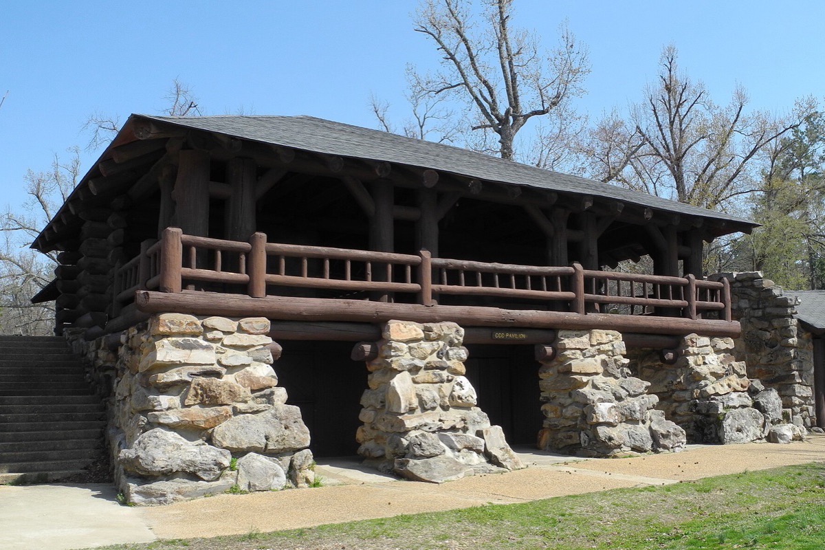 A heavy stone and timber pavilion built into a slope in Arkansas, its log railing and deep roof standing above a paved terrace with bare trees behind.