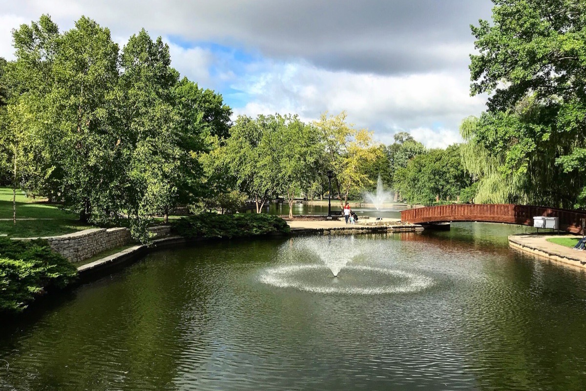 A Kansas City park pond with a fountain playing in the middle of it, a red painted footbridge at the far end and mature trees all around under scattered white cloud.