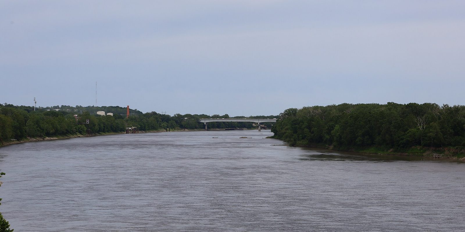Kansas City's riverfront, a wide brown river curving between dense green wooded banks toward a low highway bridge under a flat grey overcast sky