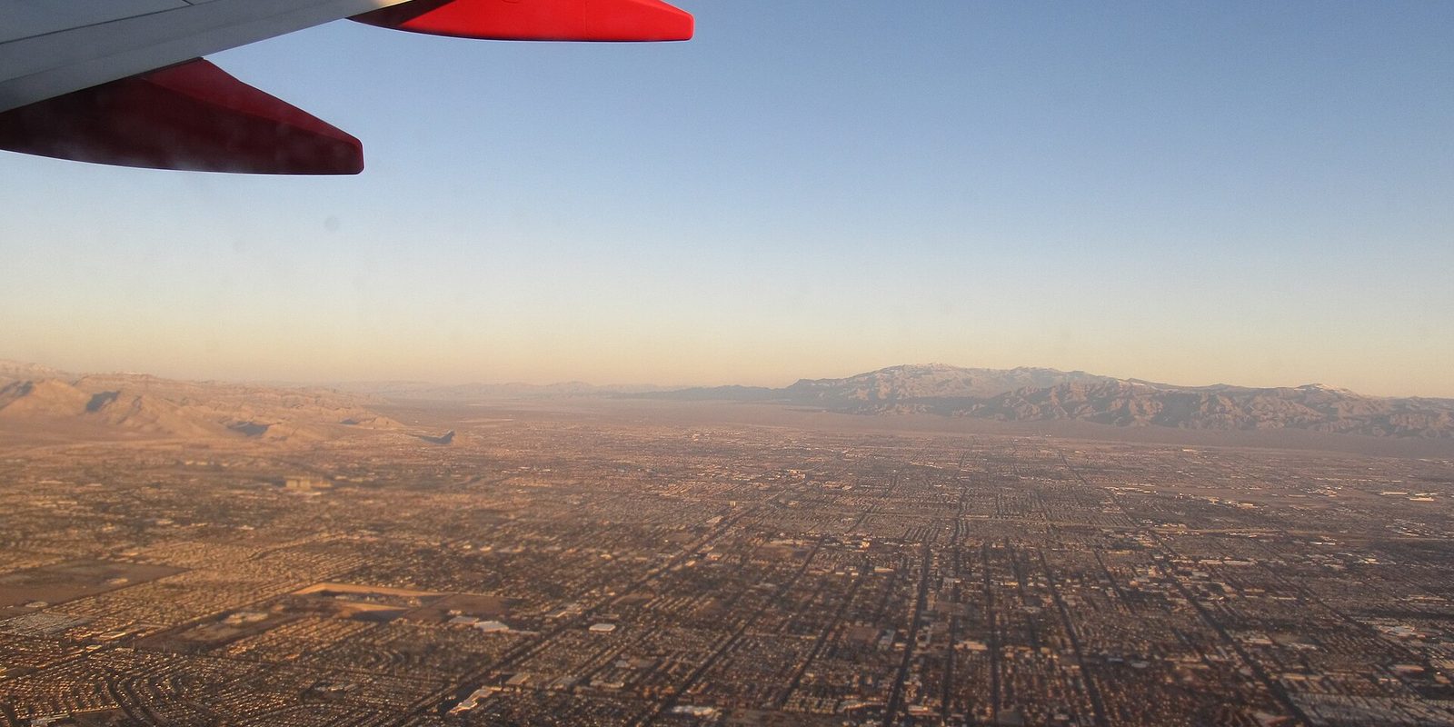 Las Vegas from an airplane window, a red wingtip above the street grid spreading across the desert valley to hazy mountains in low warm light