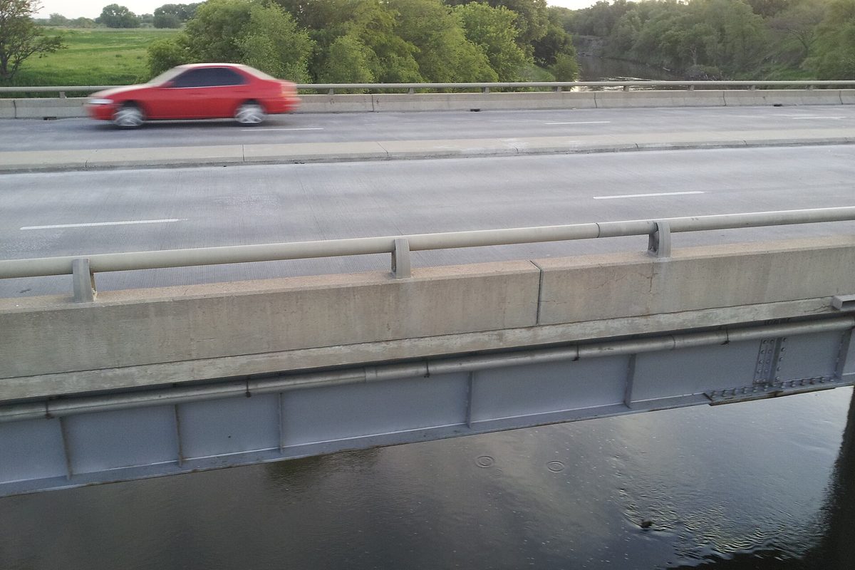 Lincoln, NE: a red sedan blurs across a concrete highway bridge over a slow river lined with leafy green trees.