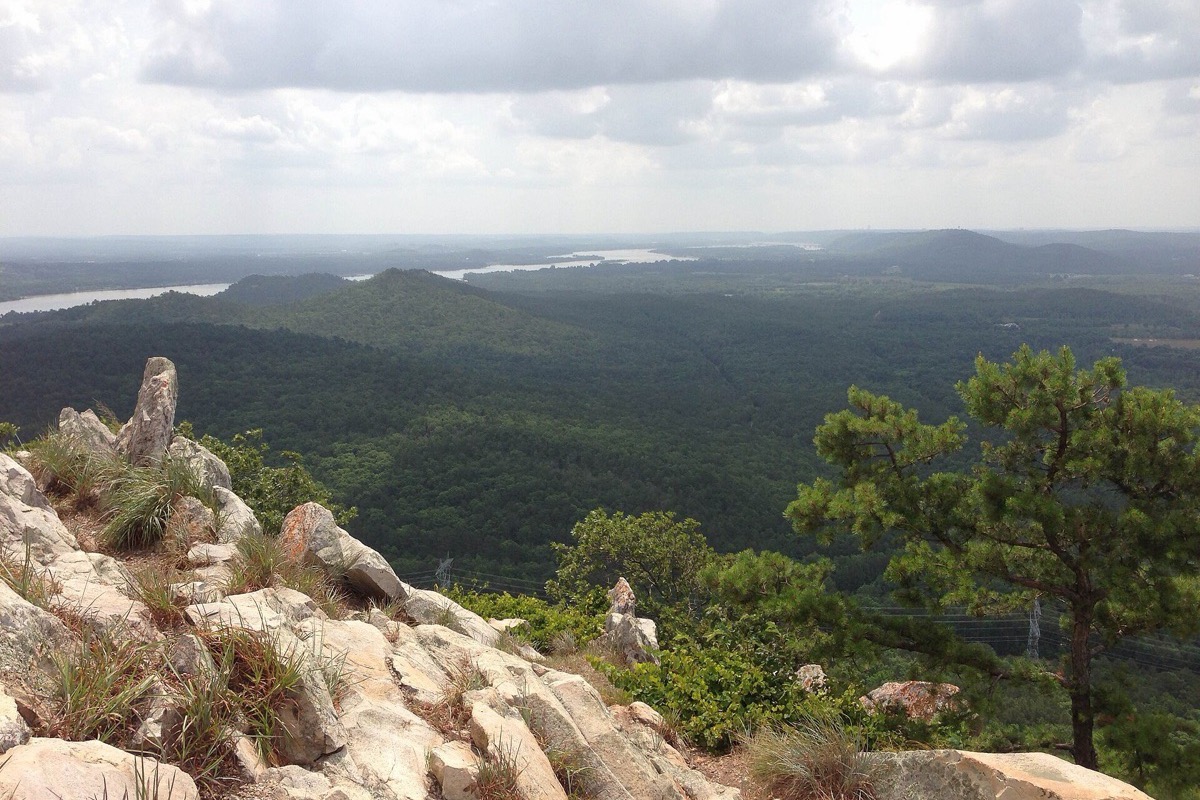 A view from a rocky Arkansas summit out over unbroken forest to a wide river valley and a line of low blue ridges on the horizon.