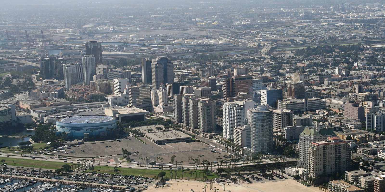 Aerial view of downtown Long Beach, California, palm-lined boulevards and high-rises between a marina and the beach, with port tanks in haze beyond