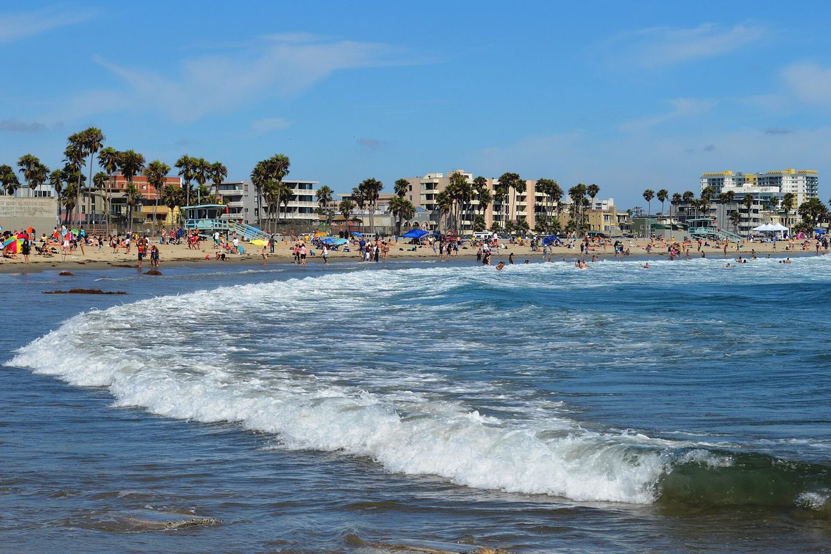Los Angeles, CA: whitewater surf rolls toward a crowded sandy beach backed by palm trees and low apartment blocks under a bright blue sky.