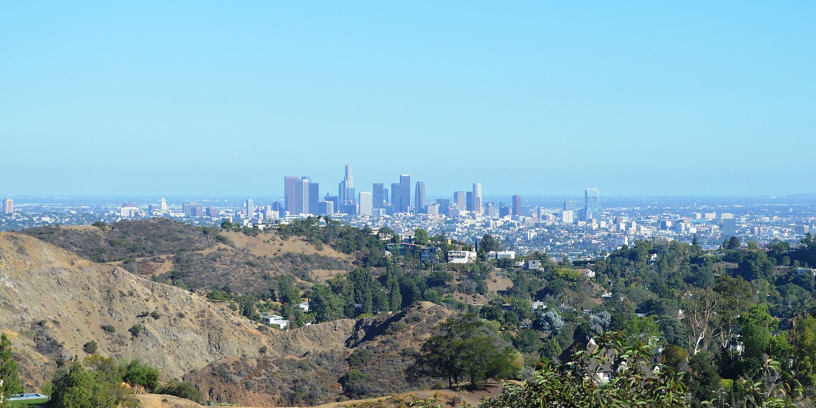 Los Angeles downtown towers on the horizon beyond dry brush-covered hills and hillside homes, under a hazy pale blue sky