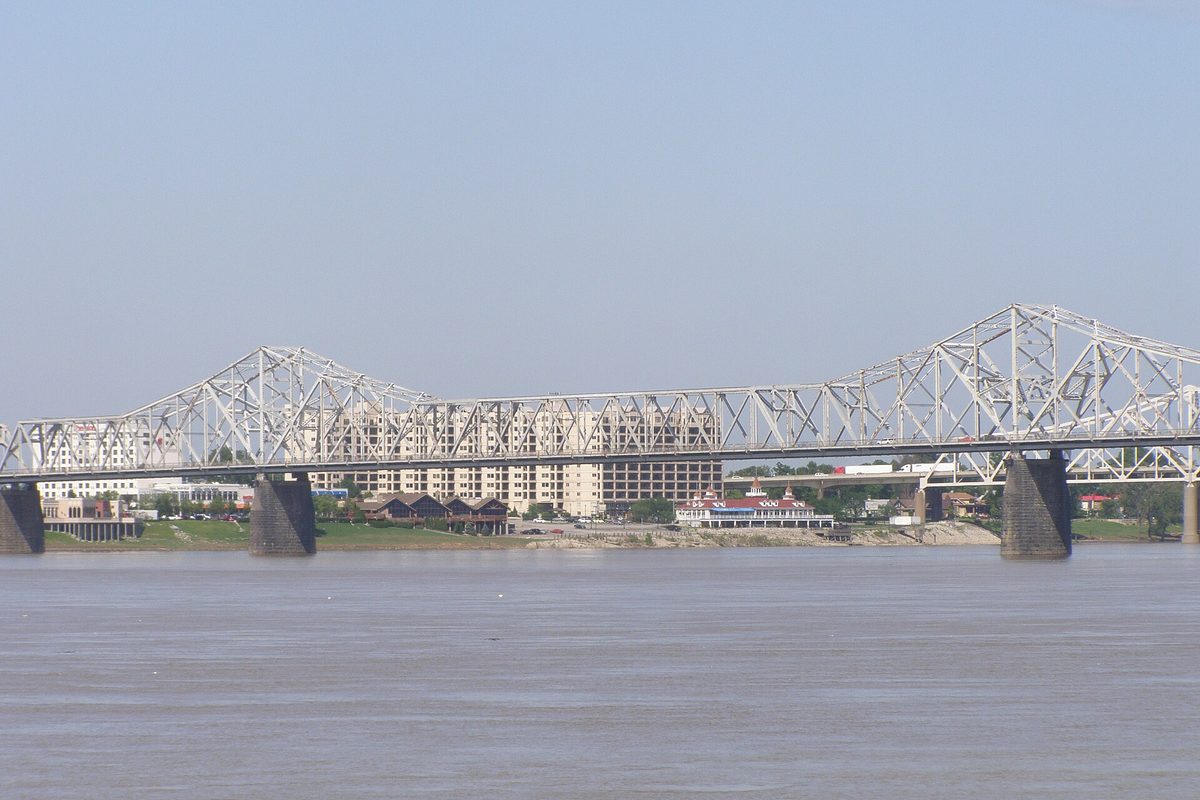 Louisville, KY: a white steel truss bridge on stone piers spans the wide brown Ohio River in front of low riverfront buildings.