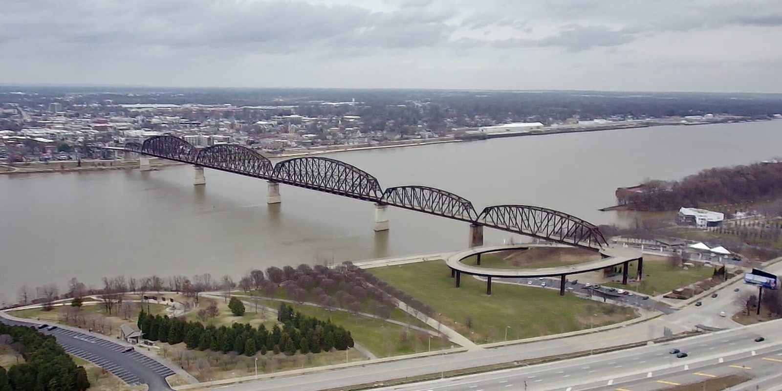 Louisville from above: a long steel truss bridge crossing the Ohio River to a riverfront park with a circular ramp, under flat grey cloud