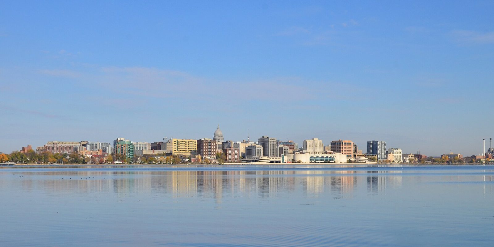 Madison's skyline and domed capitol mirrored in calm lake water under a clear blue sky, with autumn color in the shoreline trees