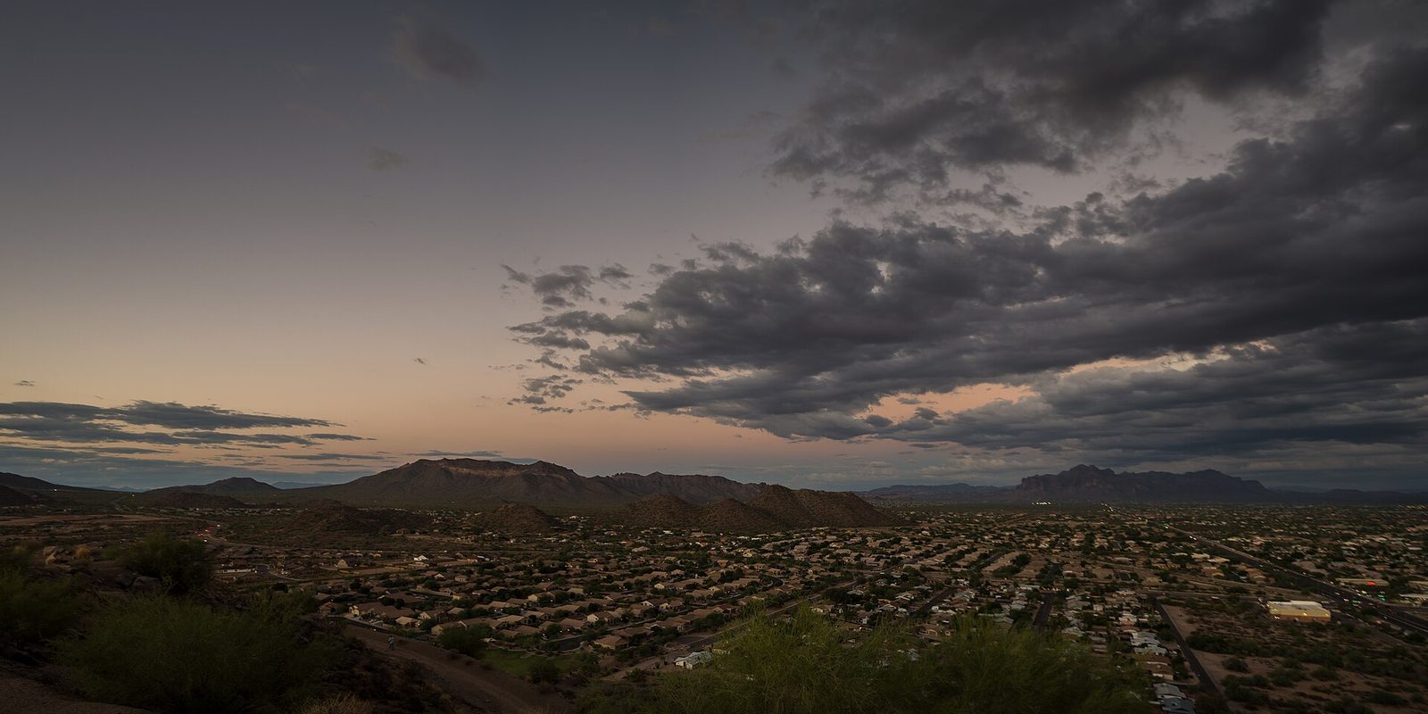 Mesa from a desert hillside at dusk, low homes spreading toward mountain ridges under heavy dark clouds and an orange band of afterglow