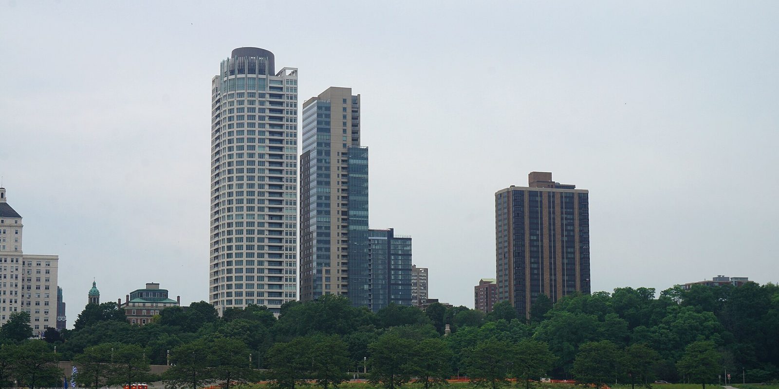 Milwaukee high-rise towers rising behind a thick band of green summer treetops in a lakefront park, under a flat pale overcast sky