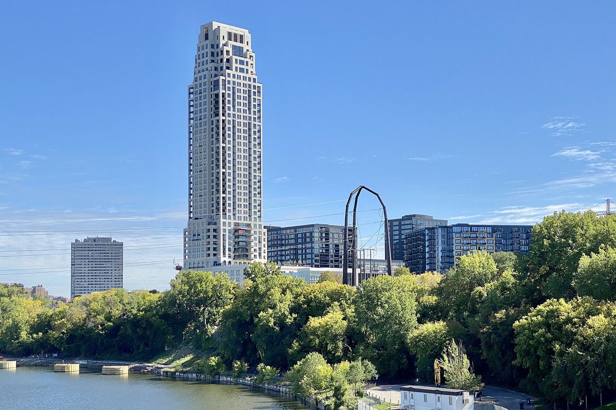 Minneapolis, a tall white condo tower and mid-rise apartment blocks above green trees on the Mississippi riverbank under a clear blue sky.