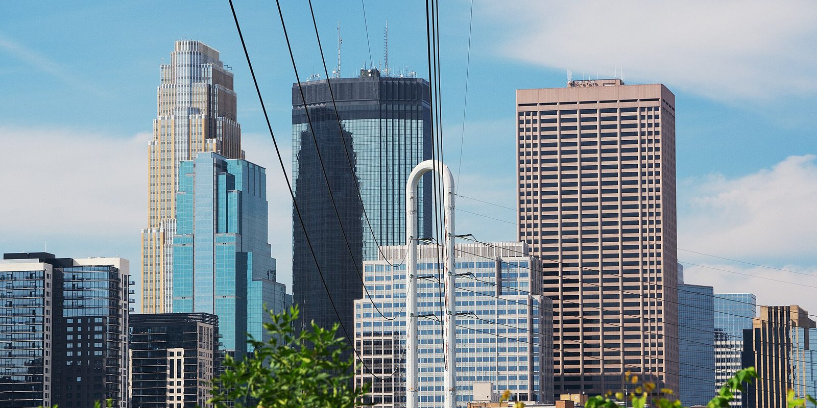 Minneapolis downtown high-rises seen close up behind overhead wires, with green leafy branches in the foreground and a blue sky